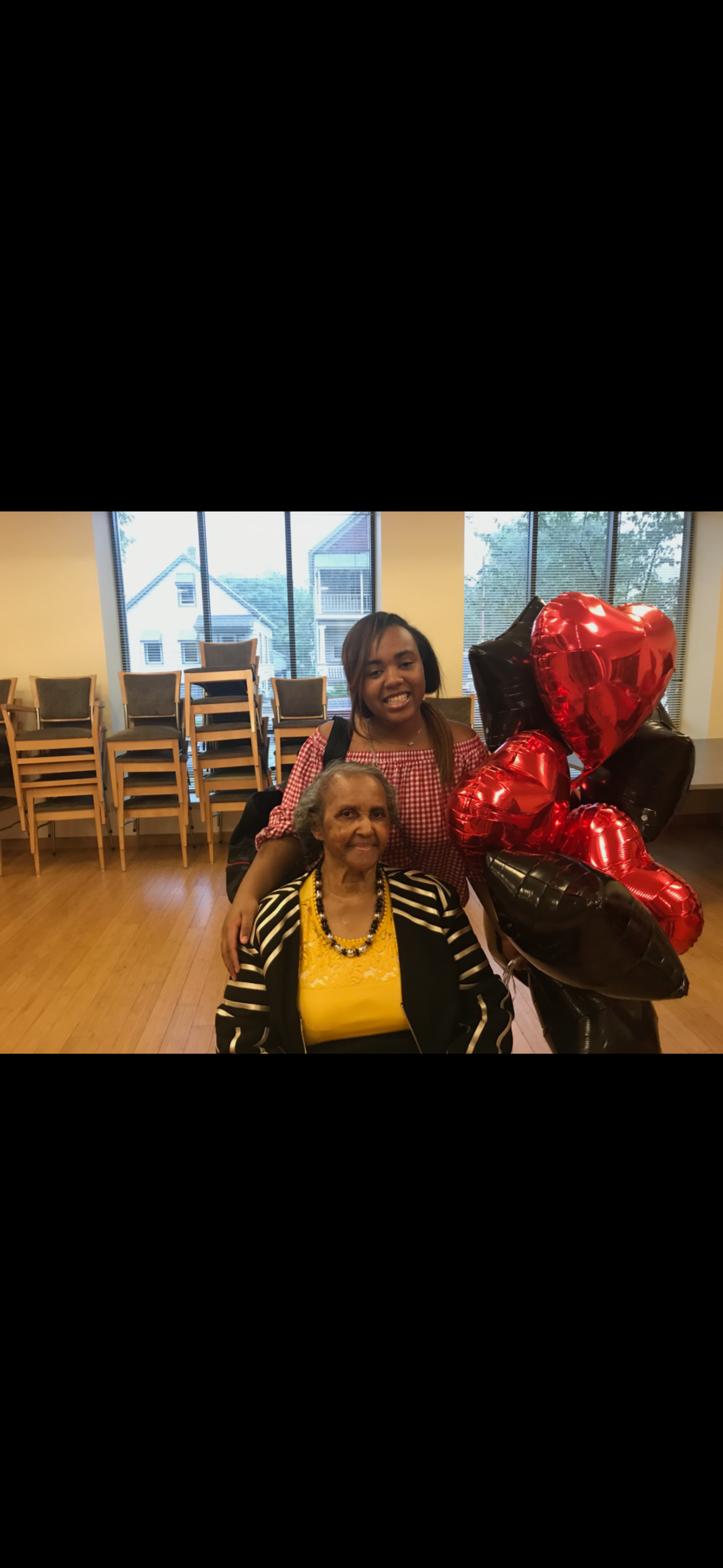 Two women pose with red and black heart balloons in a bright room; an elderly woman sits while a younger woman stands behind her smiling.