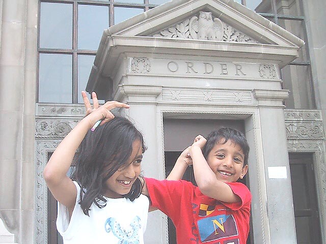 Two smiling children pose playfully with their arms raised in front of an ornate courthouse-style stone doorway.