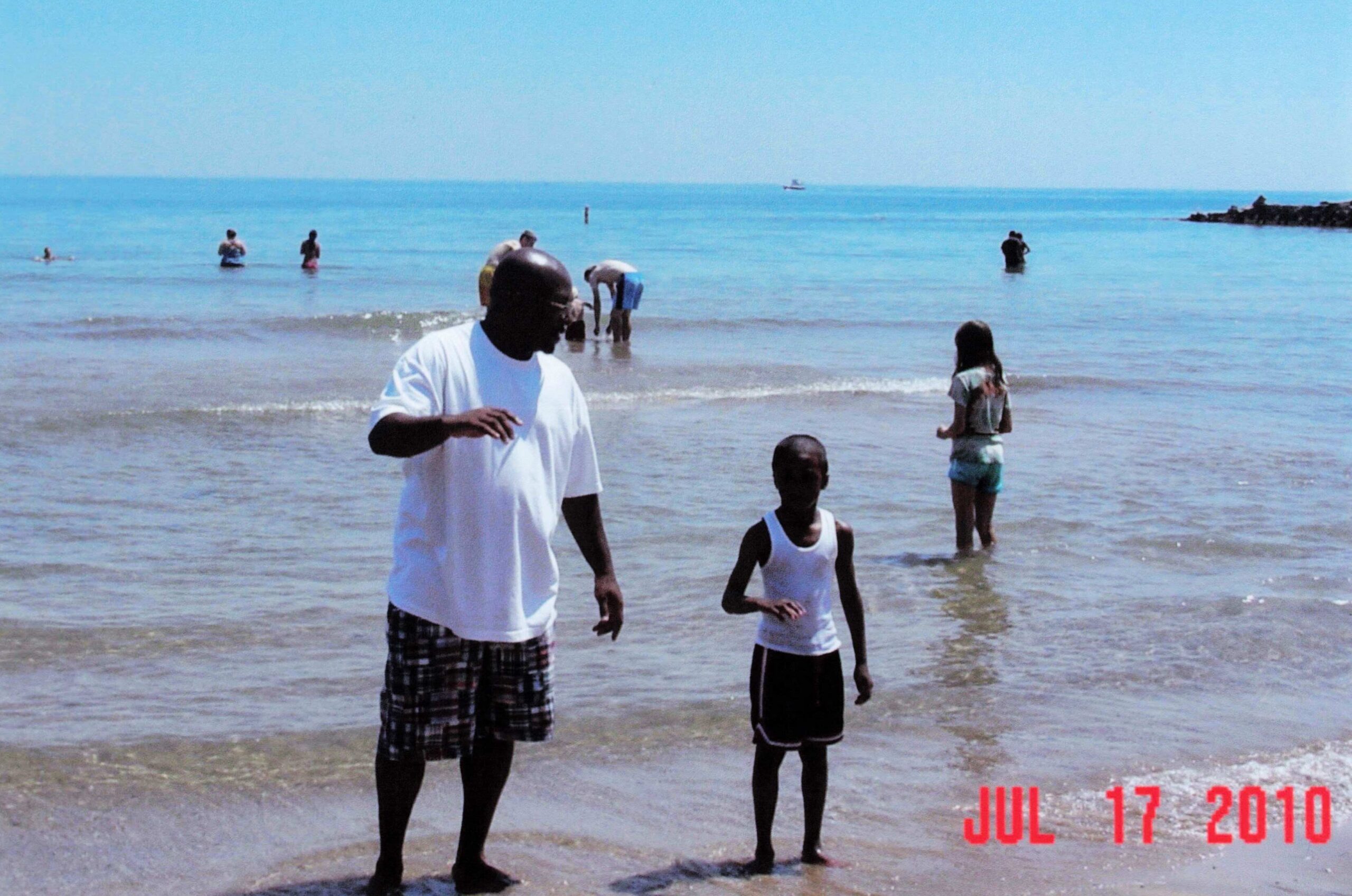 People wading in shallow ocean water at a sunny beach, with a man in a white shirt in the foreground and children nearby; calm blue sea and sky in the background. A red date stamp (Jul 17 2010) appears in the lower right corner.