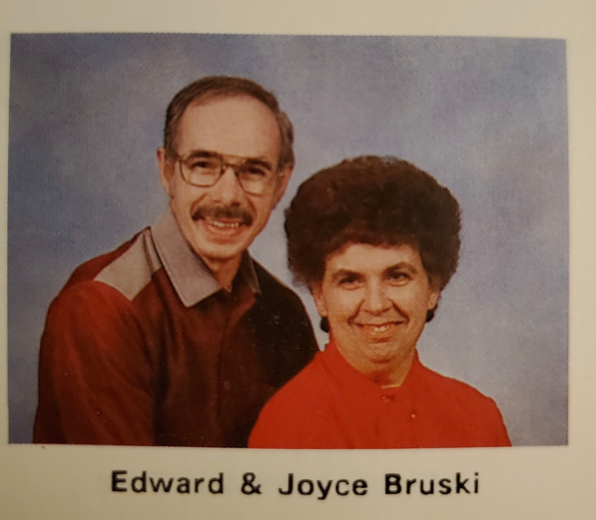 Portrait of Edward (left) and Joyce Bruski smiling together in front of a blue backdrop, Edward wearing glasses and a brown shirt, Joyce in a red top.