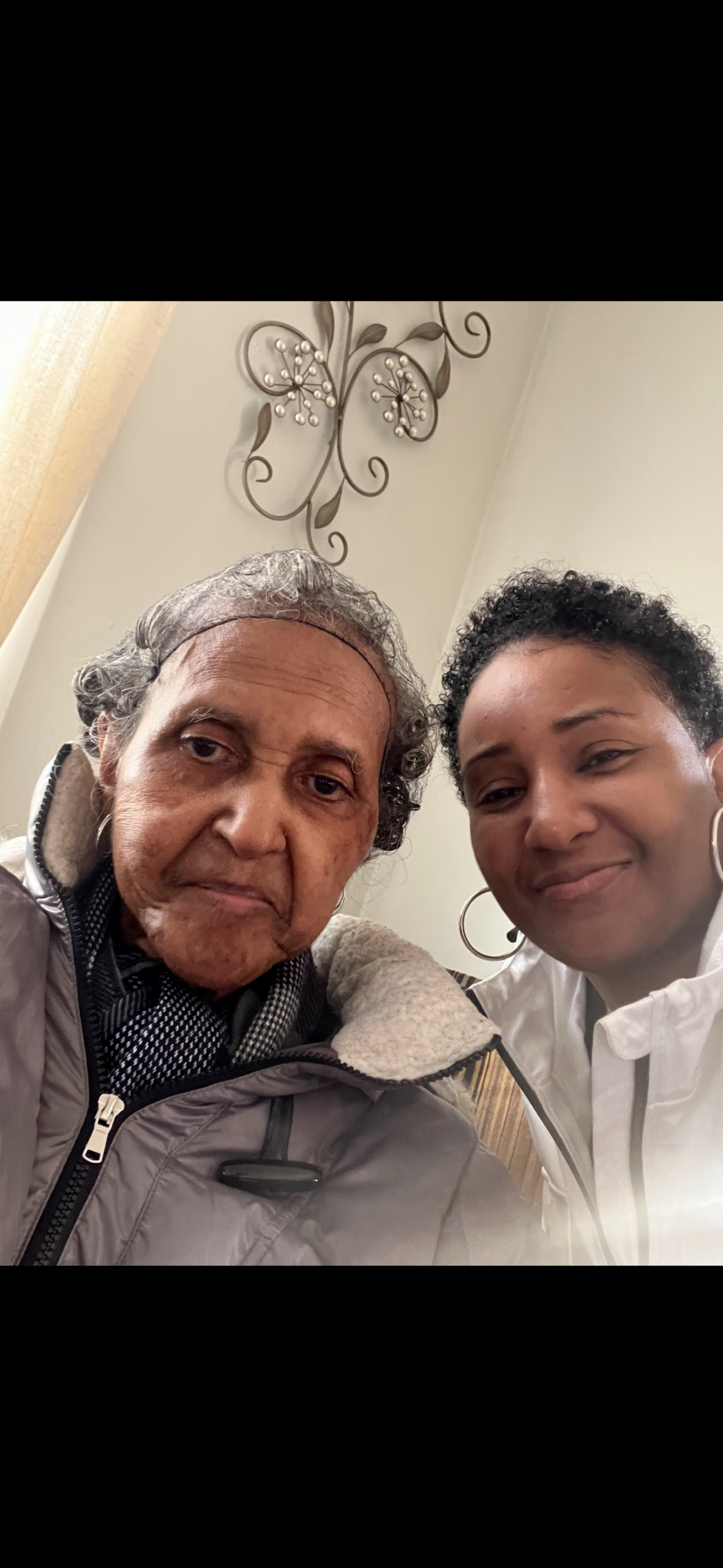 Two women taking a close-up selfie indoors, smiling at the camera with a decorative metal wall art piece in the background.
