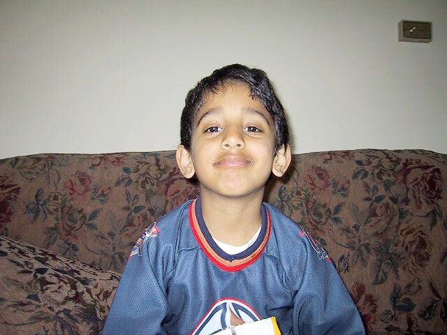 Young boy sitting on a floral-patterned sofa, smiling at the camera in a blue jersey.