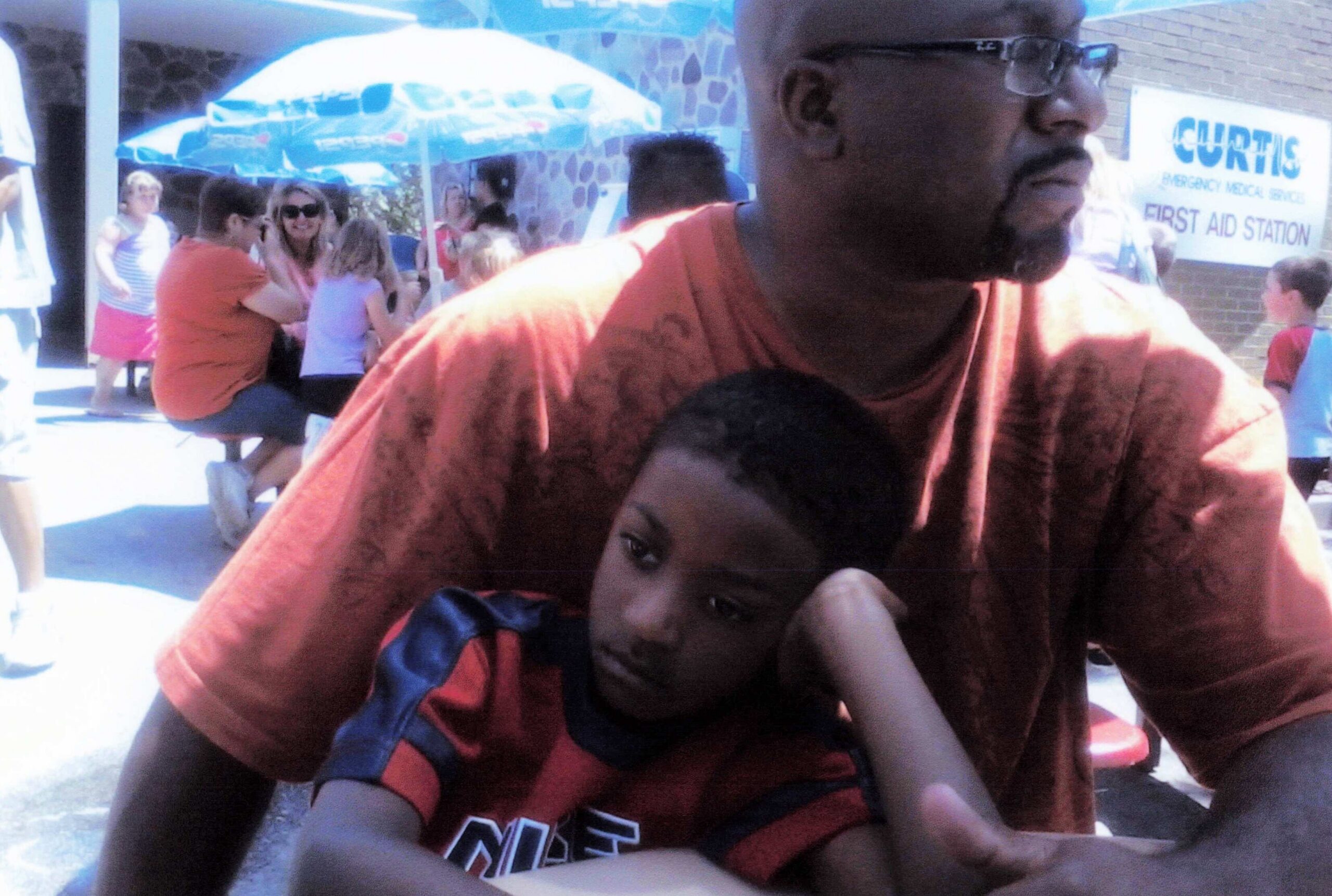 Close-up of a man in an orange shirt embracing a child wearing red and blue, outdoors at a crowded event with a first aid sign in the background.