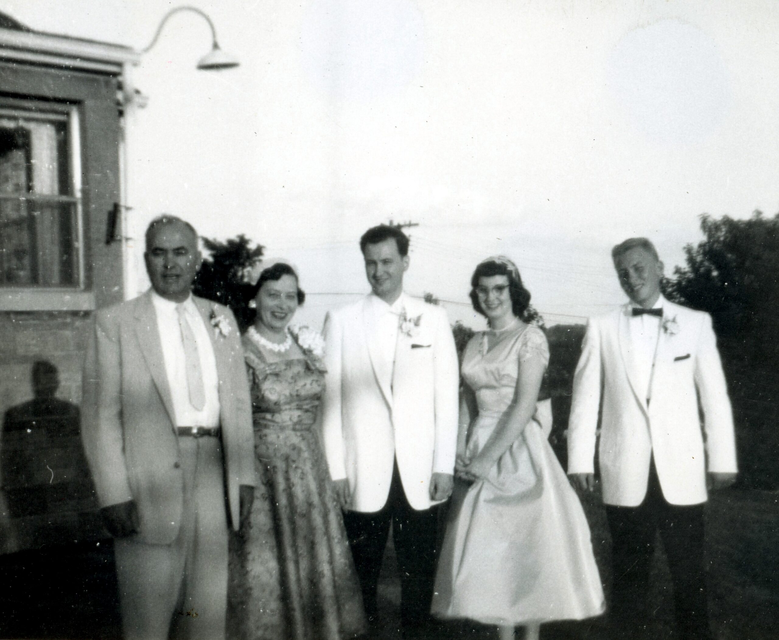 Group of six adults posing outdoors for a vintage wedding photo; three men in light suits, two women in dresses, daytime setting.