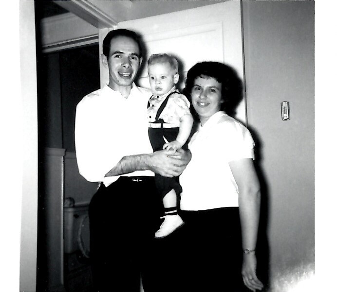 Man and woman stand with a baby in a doorway; the man holds the toddler wearing suspenders and shoes.