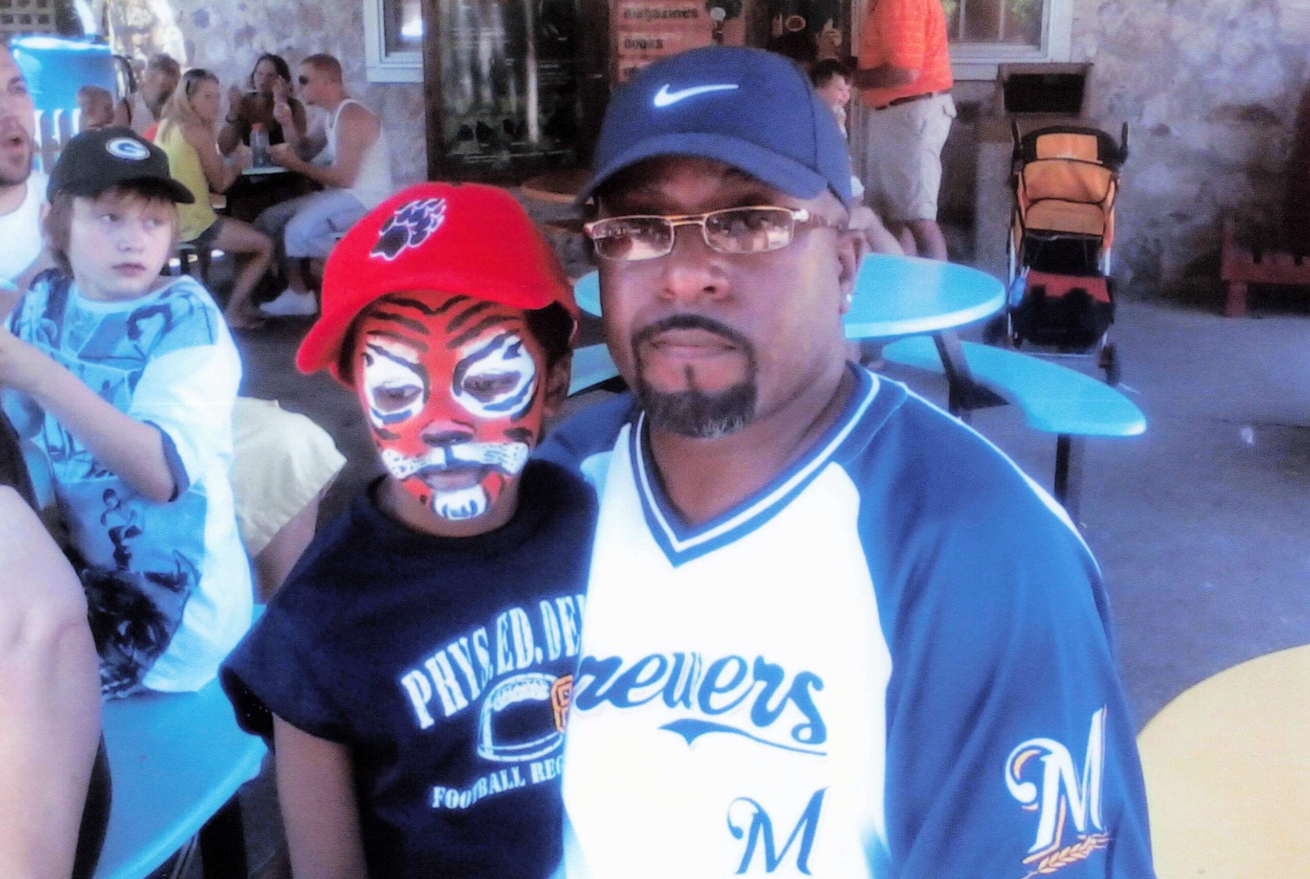 Man in a blue Nike cap and glasses taking a selfie with a child wearing tiger-face paint and a red hat at an outdoor gathering.