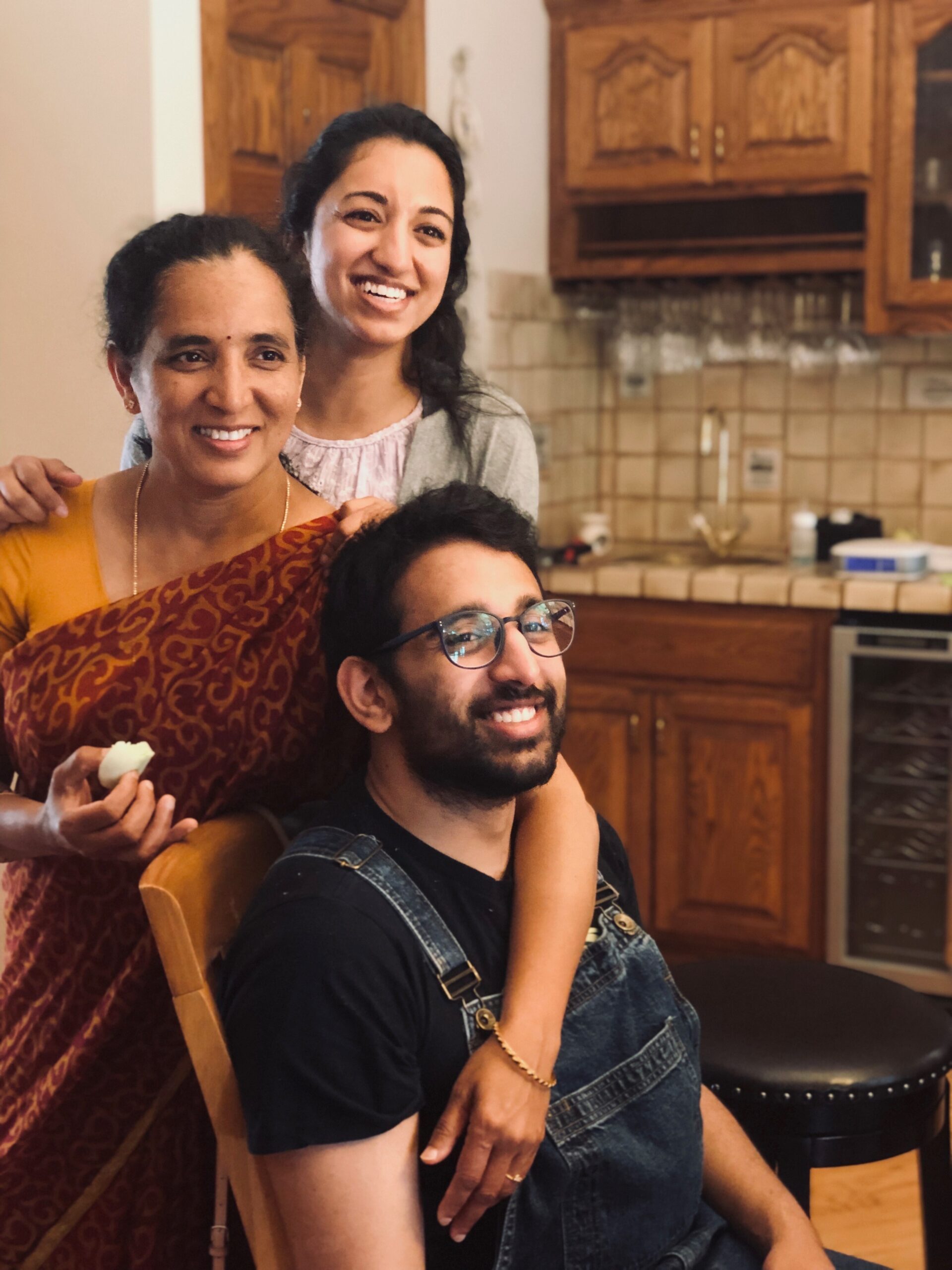 Three people posing in a home kitchen, with smiling faces; woman in an orange patterned sari in front, man in denim overalls seated, another woman stands behind them.