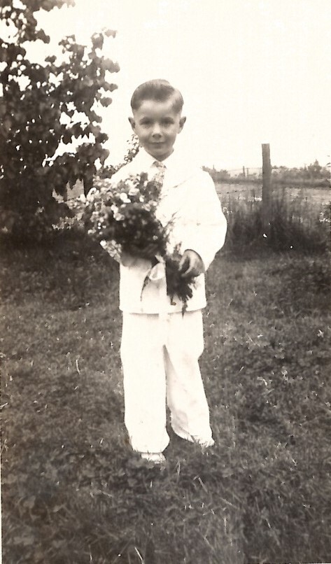 Boy in a white suit stands on grass, holding a bouquet of flowers outdoors in a garden setting.