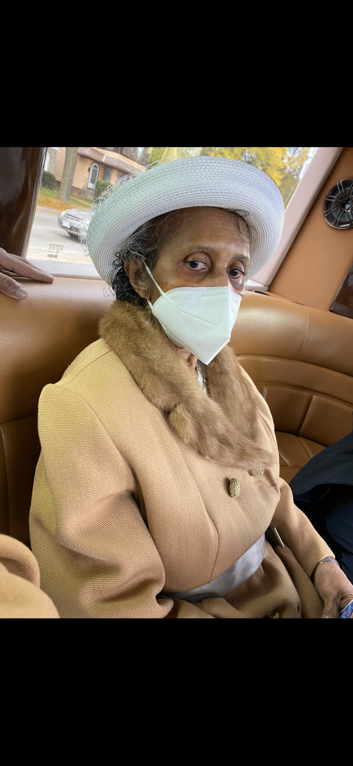 Elderly person wearing a light beige coat with fur collar and a white hat, seated in a car, wearing a mask.
