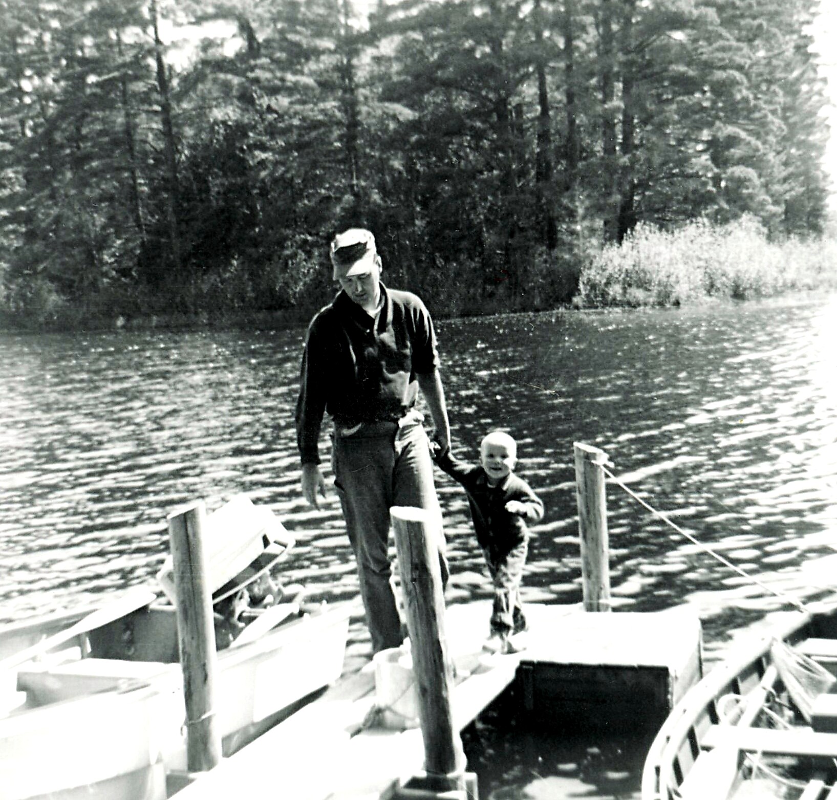 Man and young child stand on a wooden dock by a lake, with trees along the shoreline in the background.
