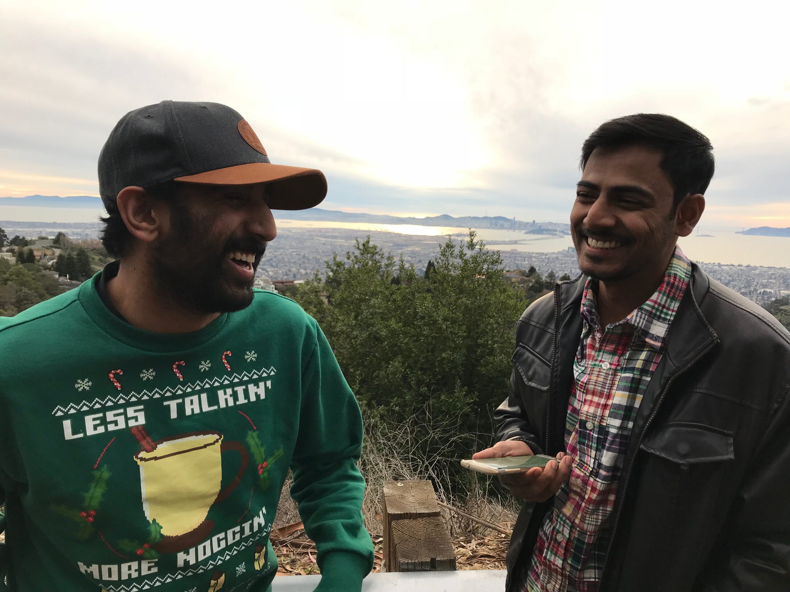 Two men smiling and talking outdoors on a hillside with a city and bay in the distance, sunset sky behind them.