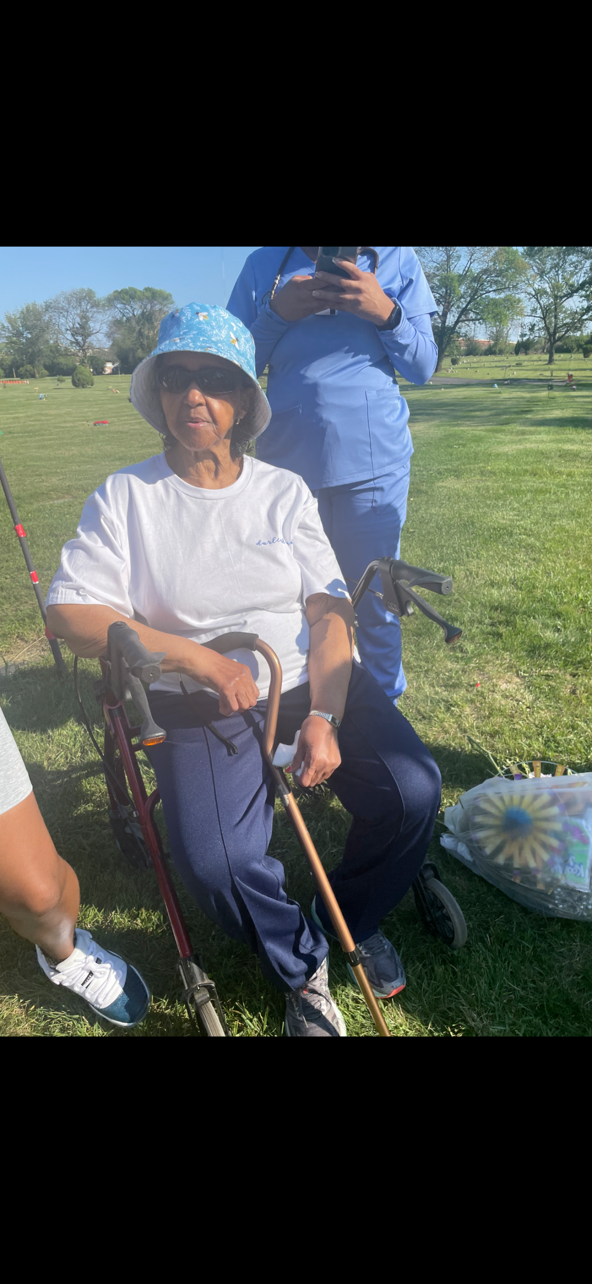 Older woman in a wheelchair outdoors in a grassy park, wearing a blue hat and white shirt, with a caregiver in blue scrubs standing behind.