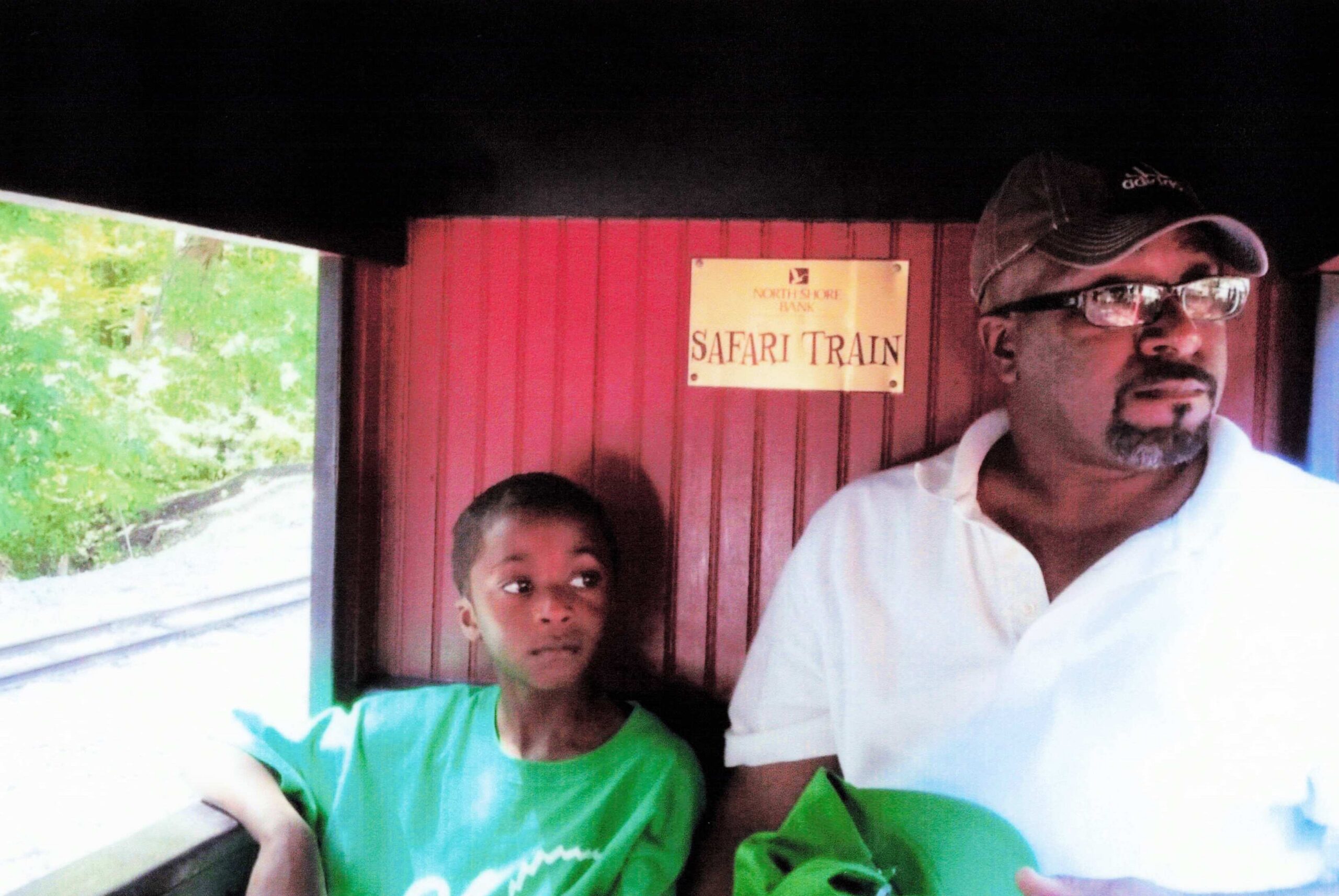 Man wearing a cap and glasses stands beside a boy in a green shirt inside a red Safari Train car.