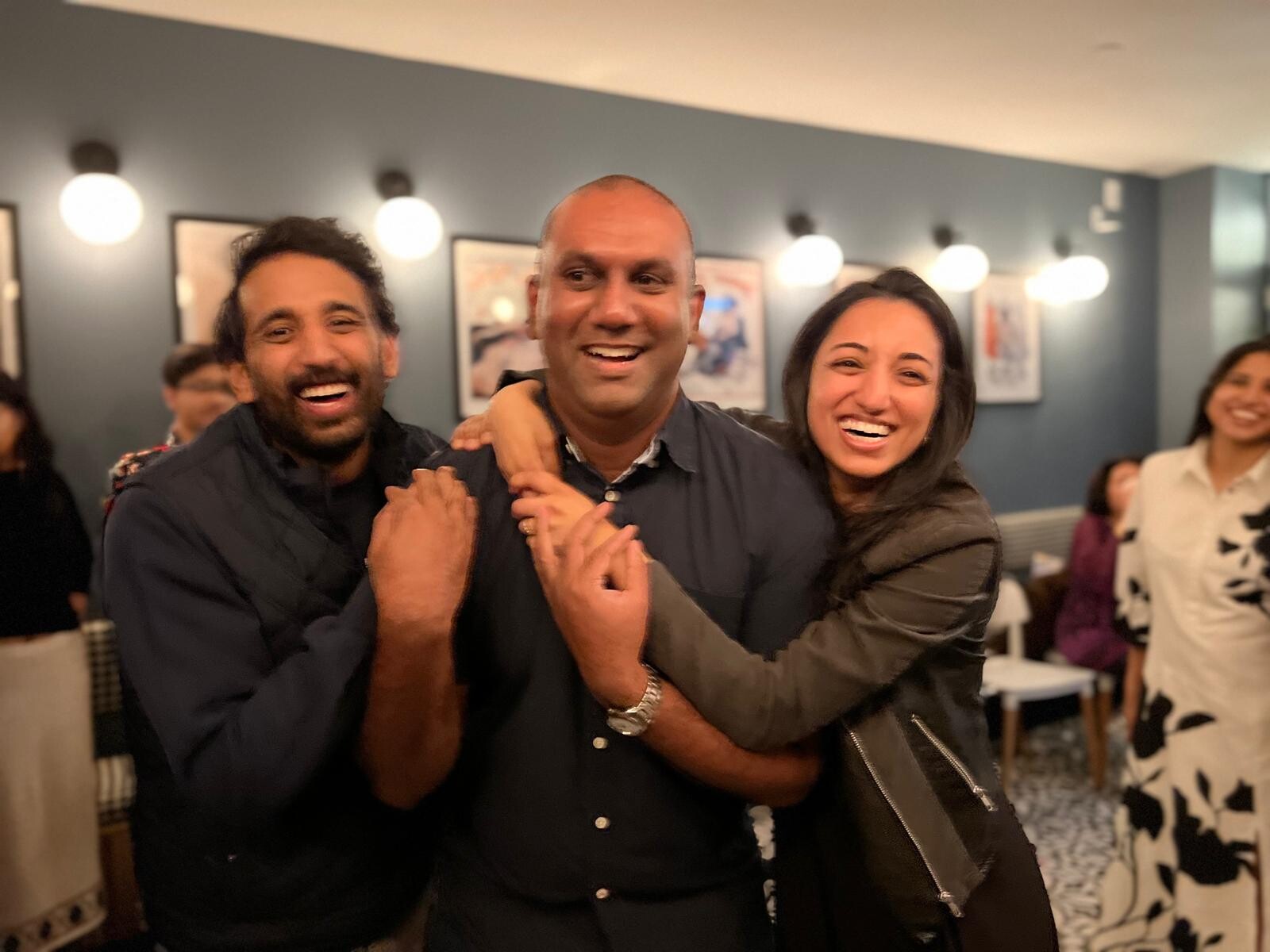 Three friends smiling and hugging at a casual indoor gathering.