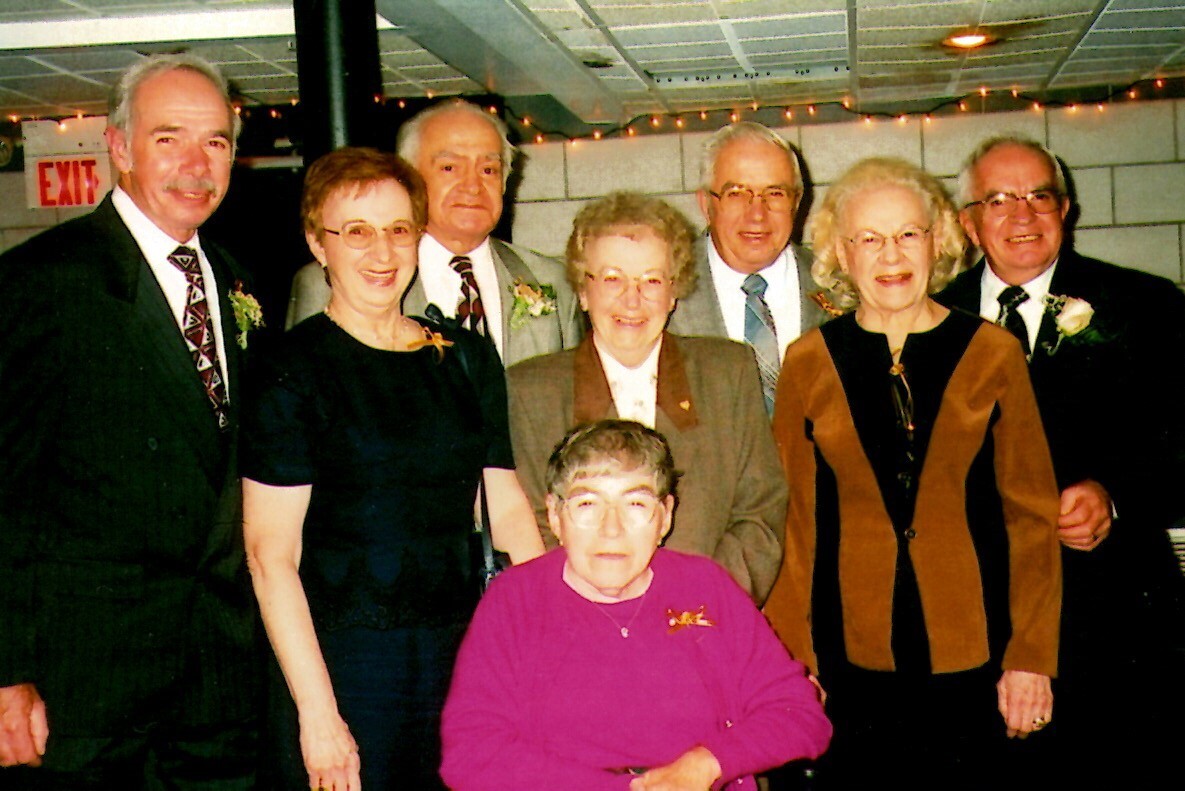 Group of eight seniors in formal attire posing for a photo at a social gathering; woman in a magenta sweater sits front center.