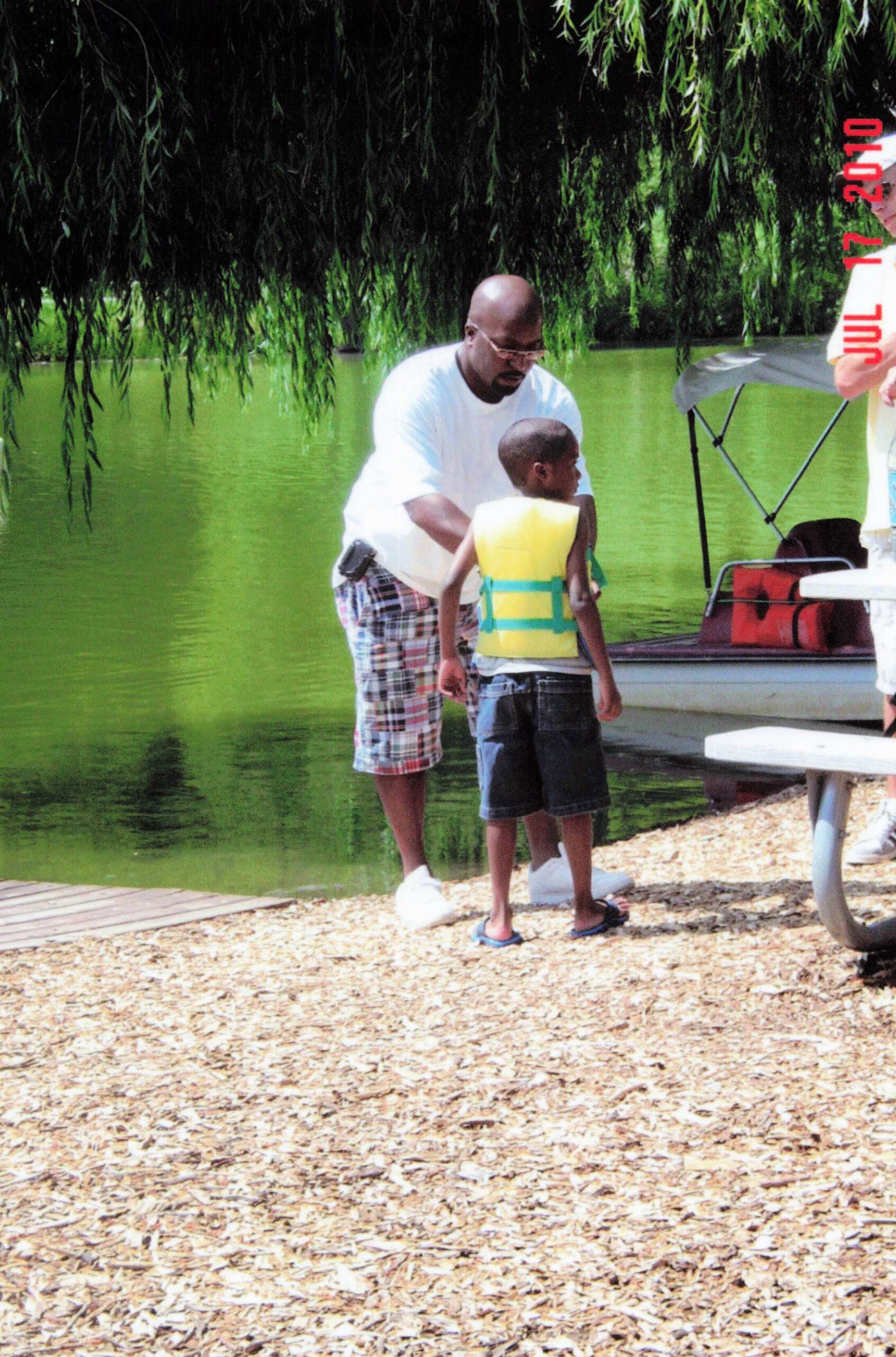 Man helping a young boy in a yellow life vest stand near a green lake, under a hanging willow tree by a docked boat.