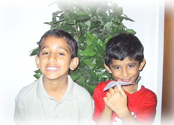 Two smiling boys pose in front of a leafy plant; the boy on the right holds a folded paper airplane near his mouth.