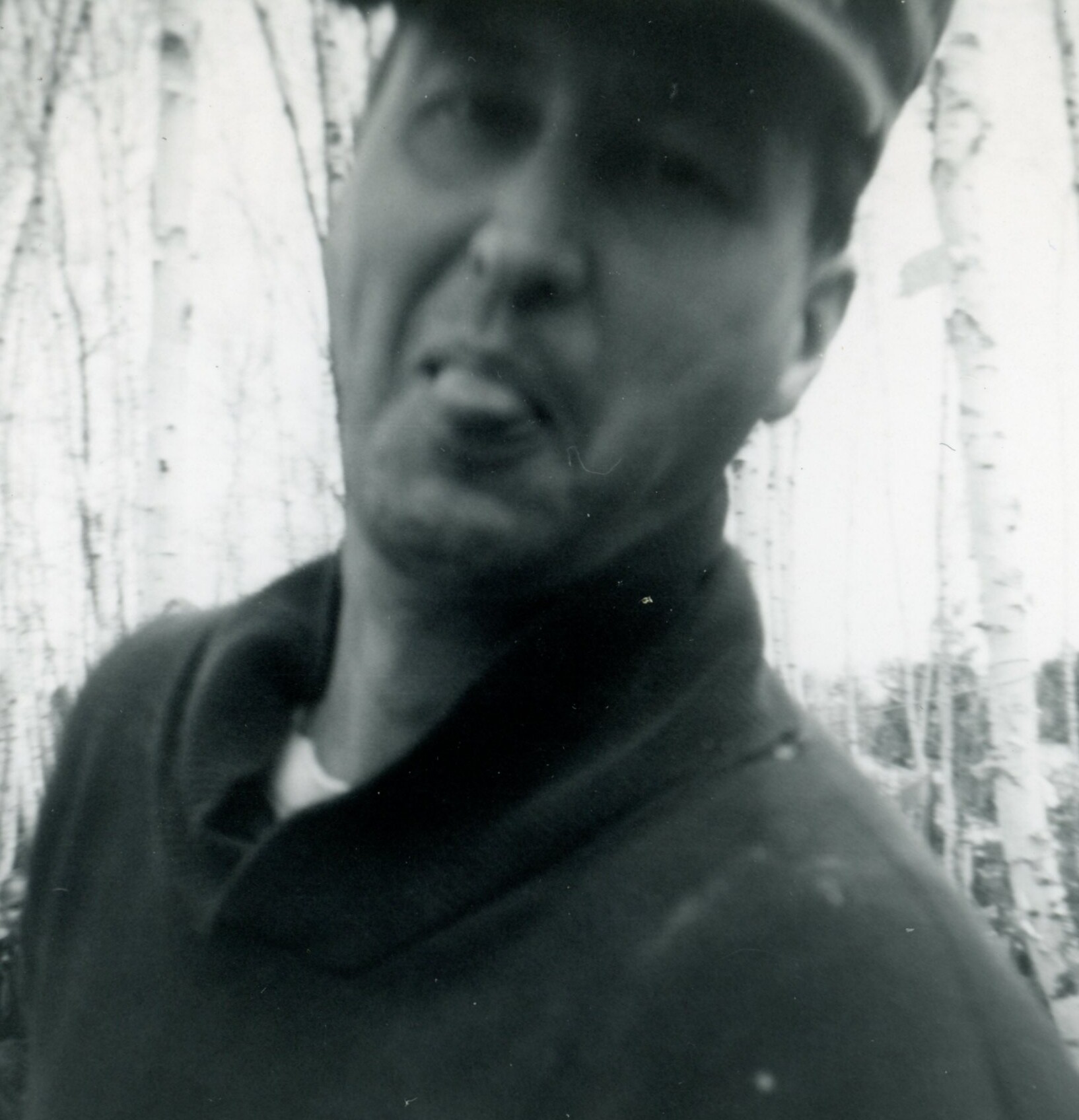 Close-up black-and-white portrait of a man outdoors, with pale birch trees in the background.