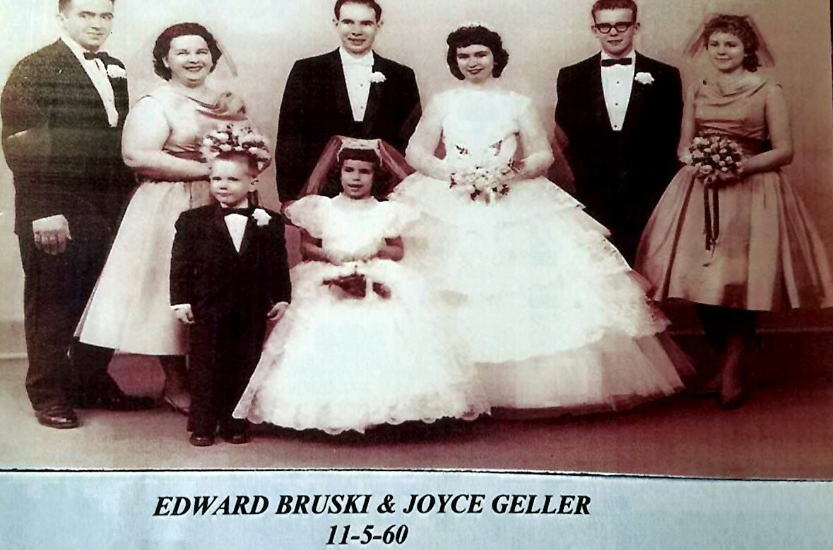 Wedding party photo: bride in a white gown and bouquet, groom in a tuxedo, flanked by bridesmaids and groomsmen in formal wear with two young children in front.
