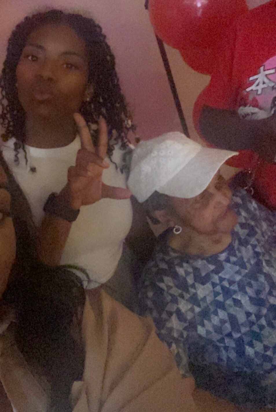 Two women posing for a selfie in a pink room; one braids wearing a white tank top shows a peace sign, the other wears a white cap and blue patterned shirt.