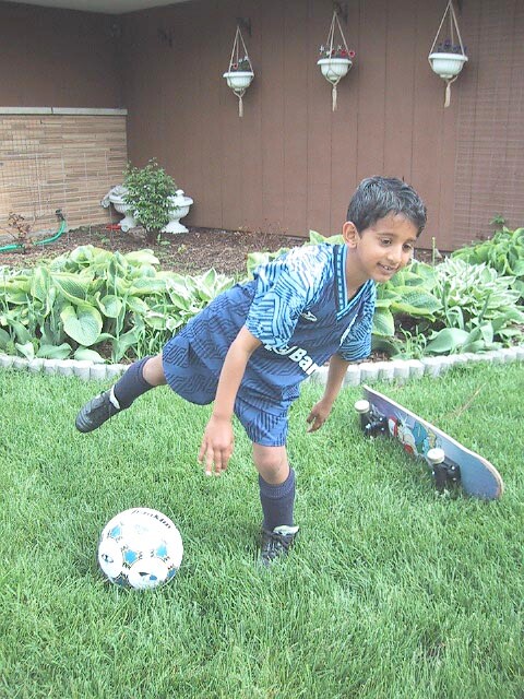 Boy in a blue soccer uniform bending to kick a ball on a green lawn, with a skateboard nearby and a garden in the background.