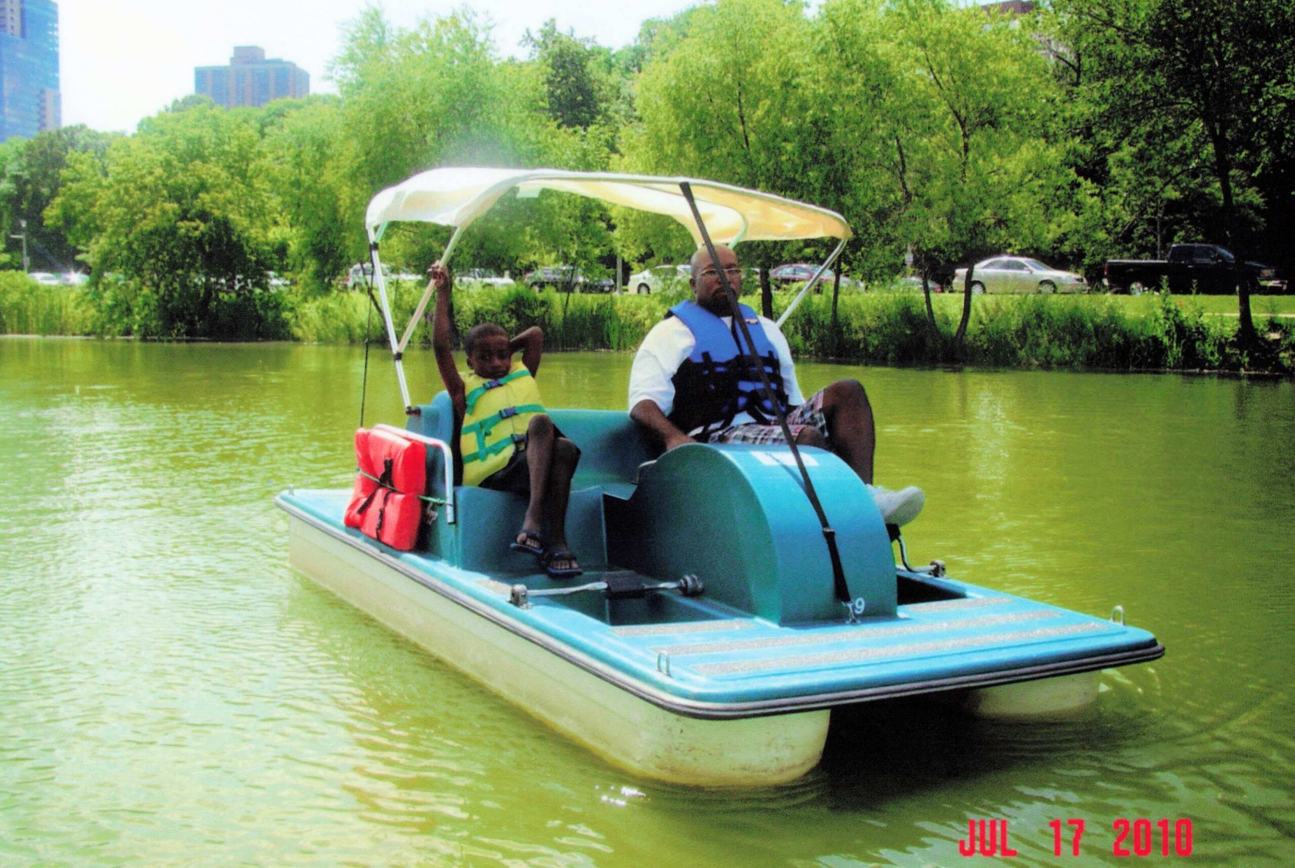 Two people in a blue pedal boat wearing life jackets glide on a greenish lake with trees and distant buildings in the background.