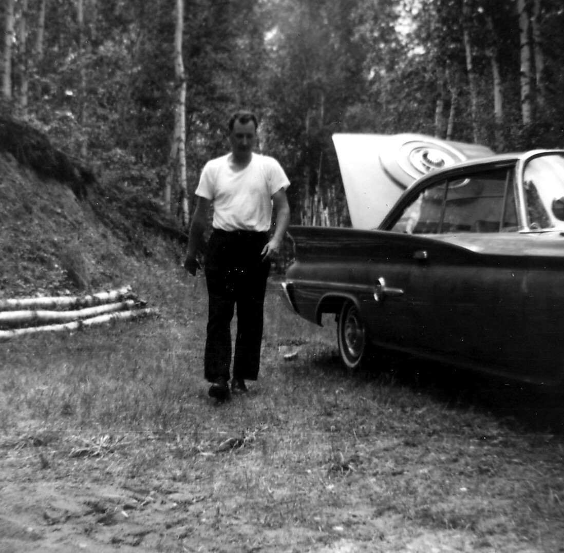 Man in a white T-shirt and dark pants walks along a dirt path beside a vintage car with its hood raised in a wooded area.