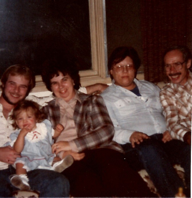 Family sits on a sofa: a man with a toddler girl on his lap, flanked by two women and another man smiling up at the camera.