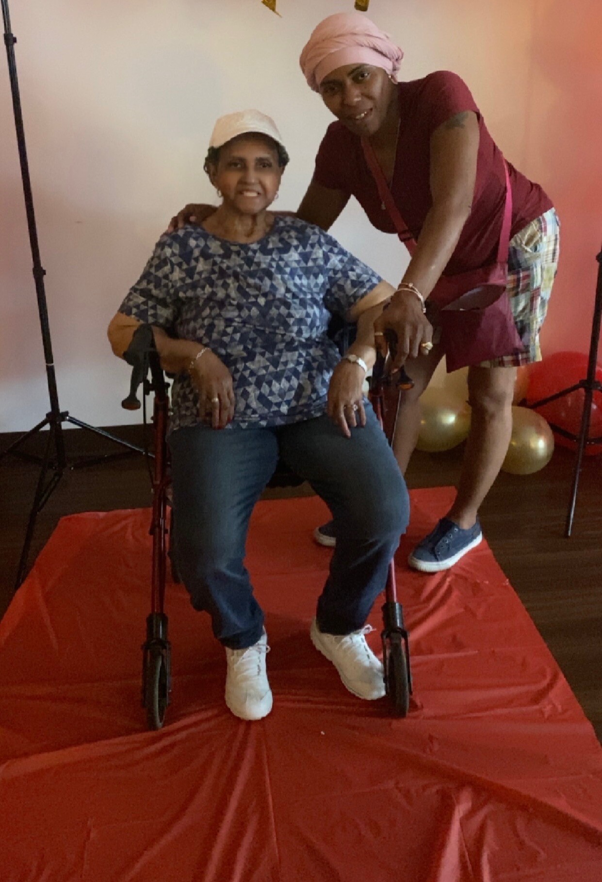 Two women pose for a photo in a home studio; an elderly woman sits in a walker with a blue geometric blouse, white cap, and white sneakers, while a caregiver in a maroon top and pink head wrap stands beside her.