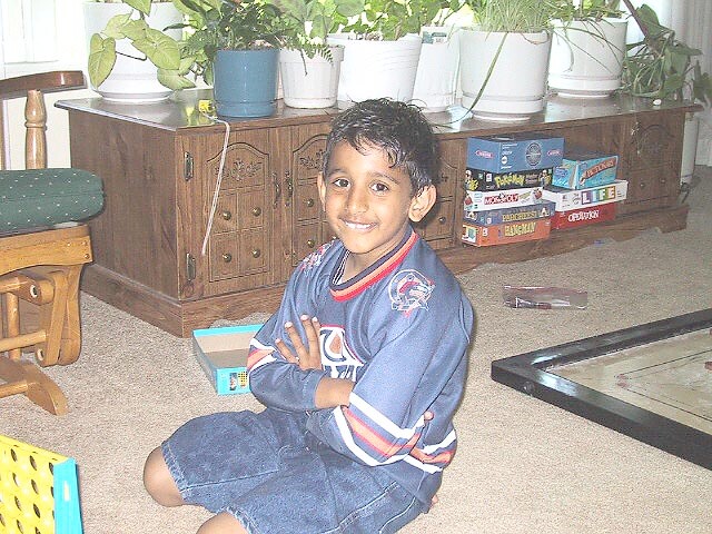 Smiling young boy sitting cross-legged on a carpet in a living room, wearing a blue hockey jersey, with plants and board games on a cabinet behind him.