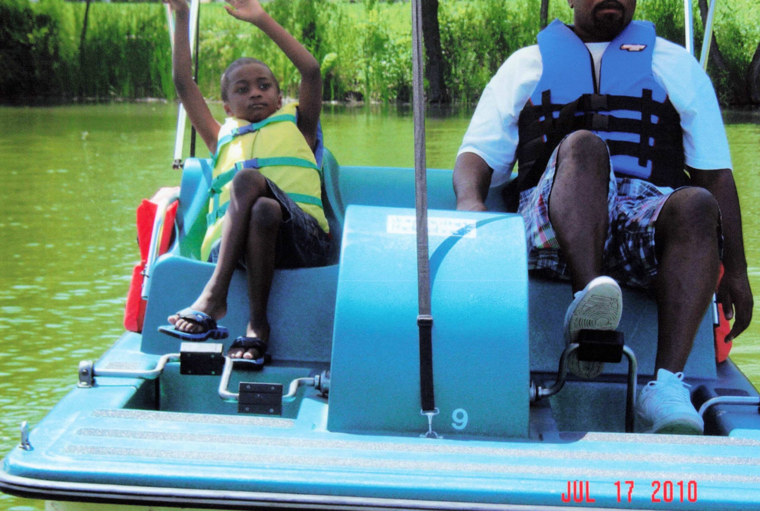 Child and adult in a blue boat on a green lake, both wearing life jackets; the child raises arms in the front left.