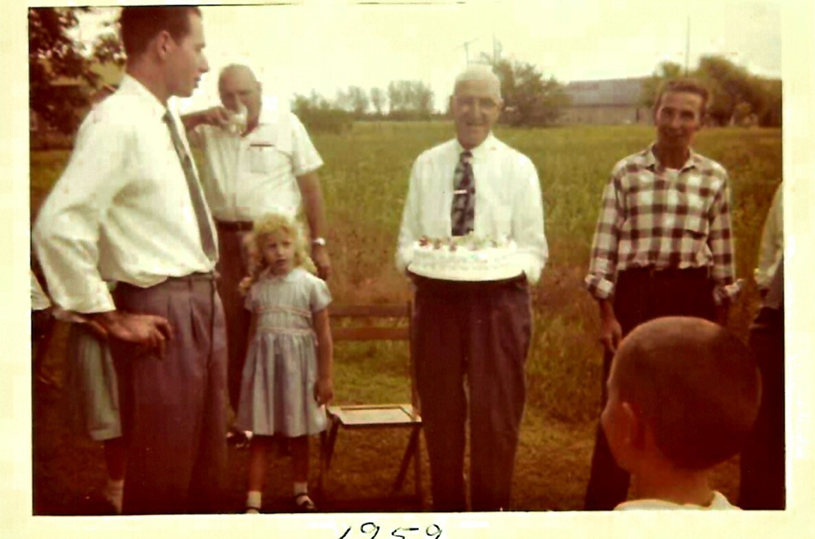 Group of adults and a young girl outdoors for a birthday, with an older man in a white shirt and tie holding a cake.