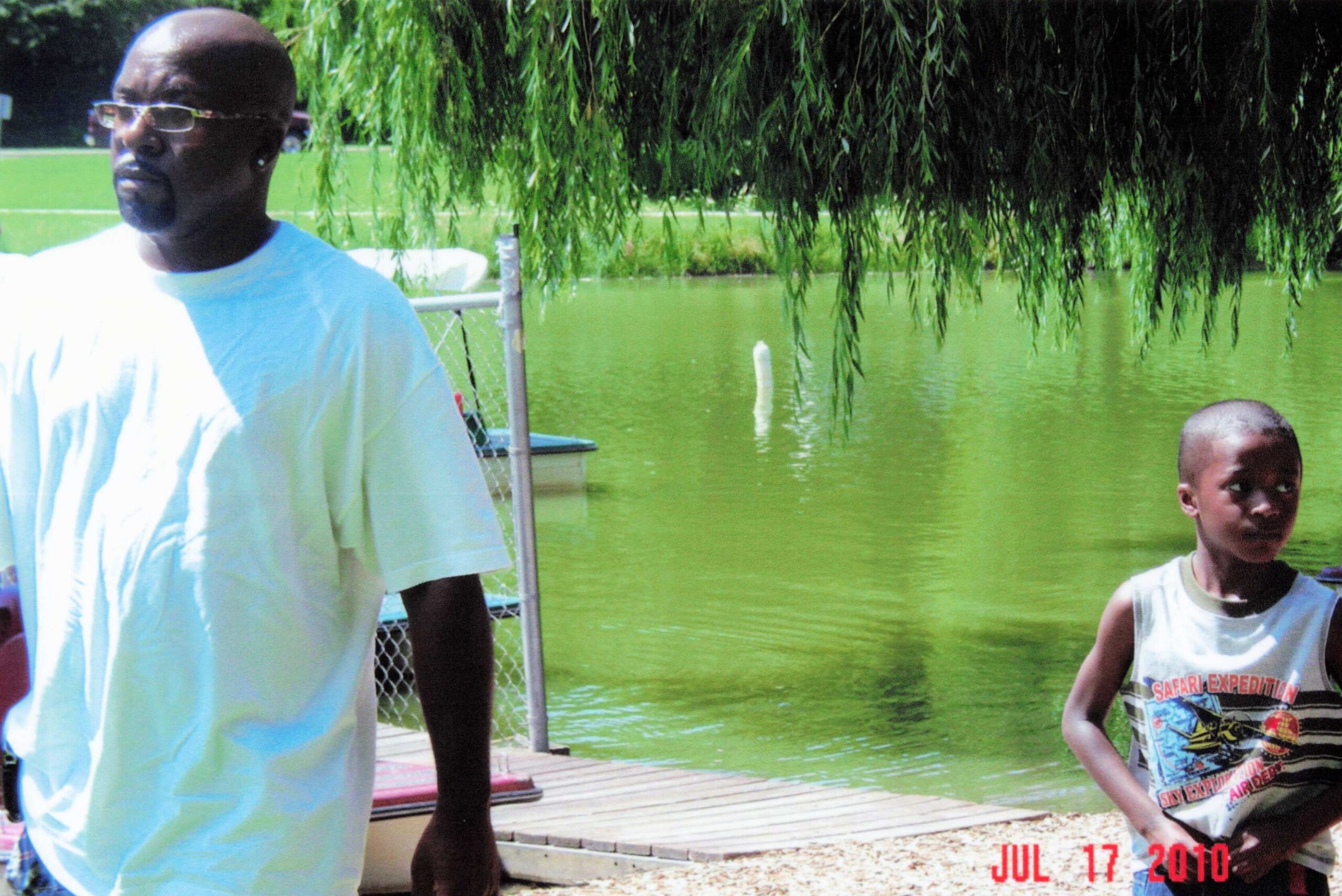 Two people stand on a wooden dock beside a green pond under a willow tree; man in a light blue shirt and glasses, boy in a white sleeveless shirt.