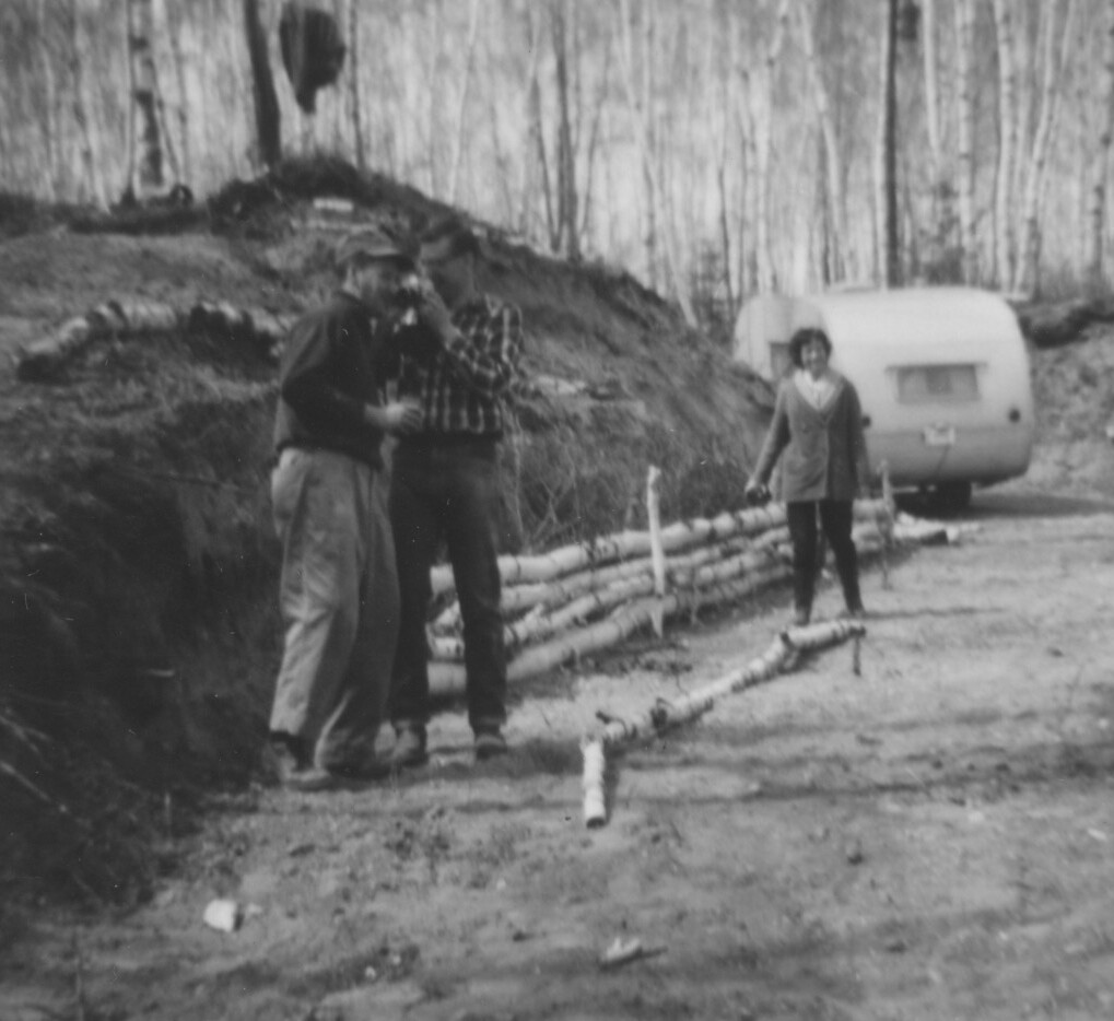 Black-and-white photo of two men and a woman on a dirt road in a forest; a small caravan sits in the background.