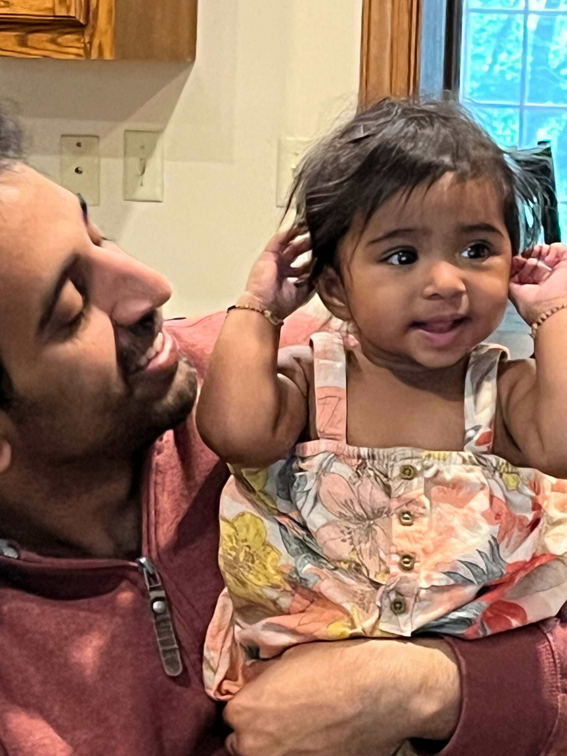 Father smiles at a toddler girl as she touches her hair in a home setting, likely a kitchen area behind them.