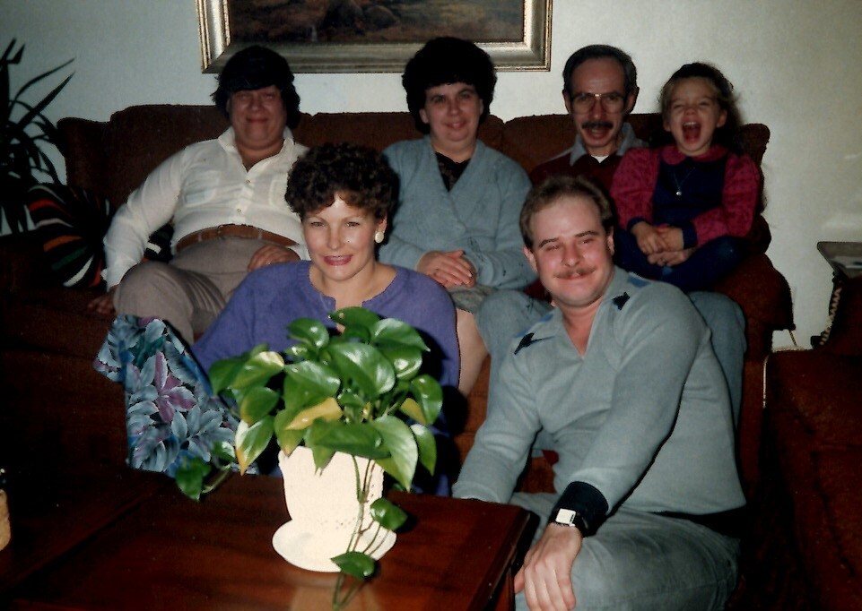 Family group portrait in a living room: six relatives seated on a couch and chair, with a woman in a purple top and a man in a gray sweater in the foreground, a child laughing on the right, and a potted plant on a coffee table in front; framed painting on the wall behind them.