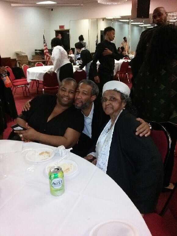 Three people at a round banquet table pose for a photo, smiling and hugging, with a can of soda in the foreground.