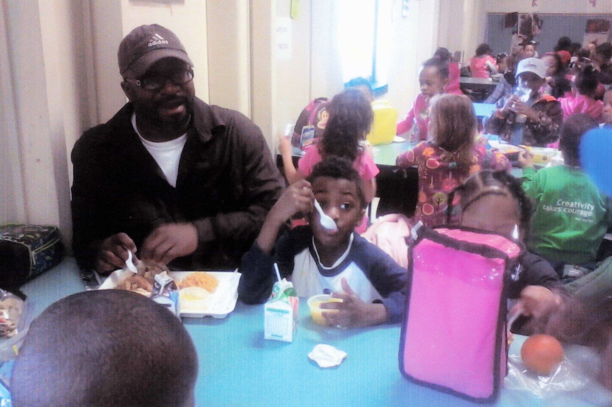 A man wearing a dark cap sits beside several children at a colorful table, eating and chatting during a crowded event.