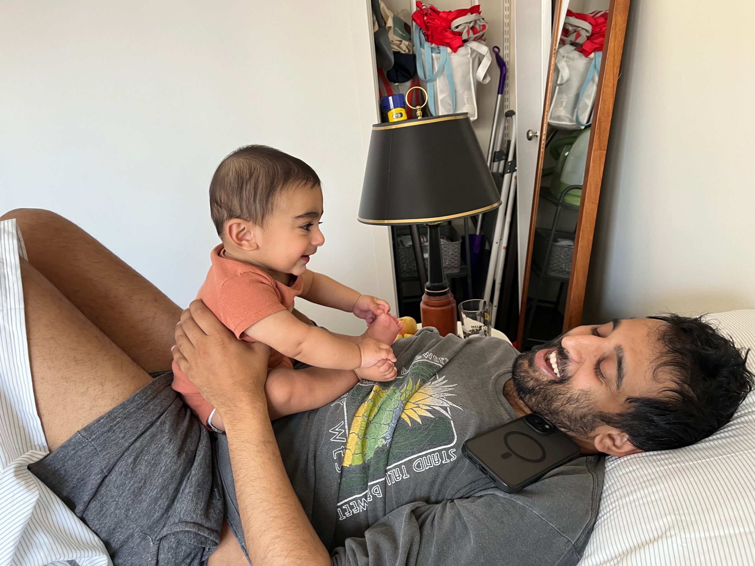 Bearded man lying in bed, smiling, while holding a baby wearing an orange shirt; a lamp and closet with a mirror visible in the background.