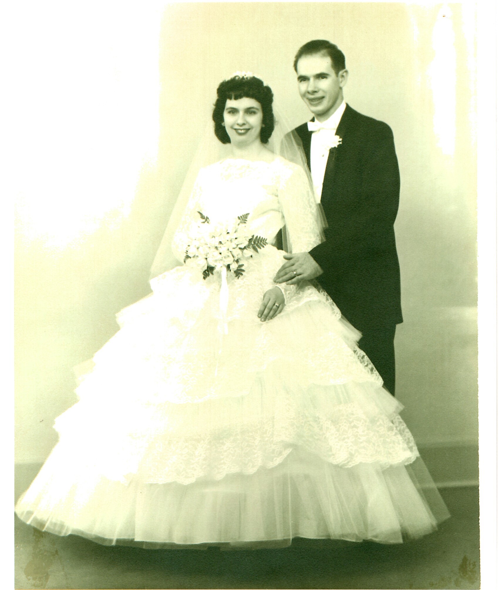 Bride in a lace wedding gown holding a bouquet, standing beside a man in a black tuxedo (the groom) in a studio portrait.