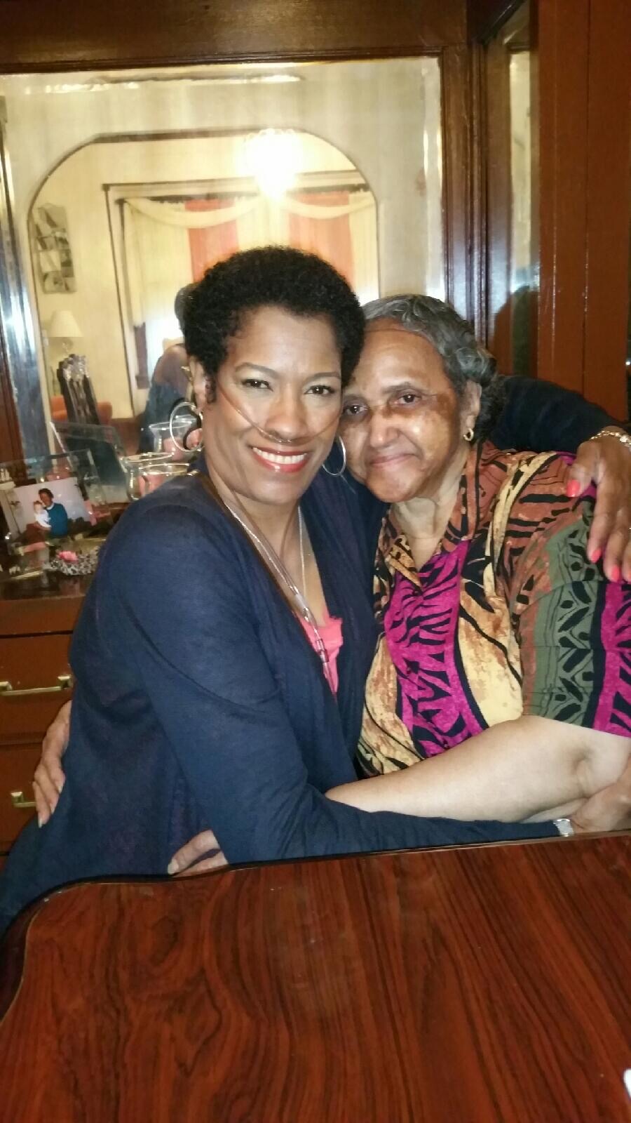 Two women smile and hug at a wooden table in a warm room; one has nasal oxygen tubing.