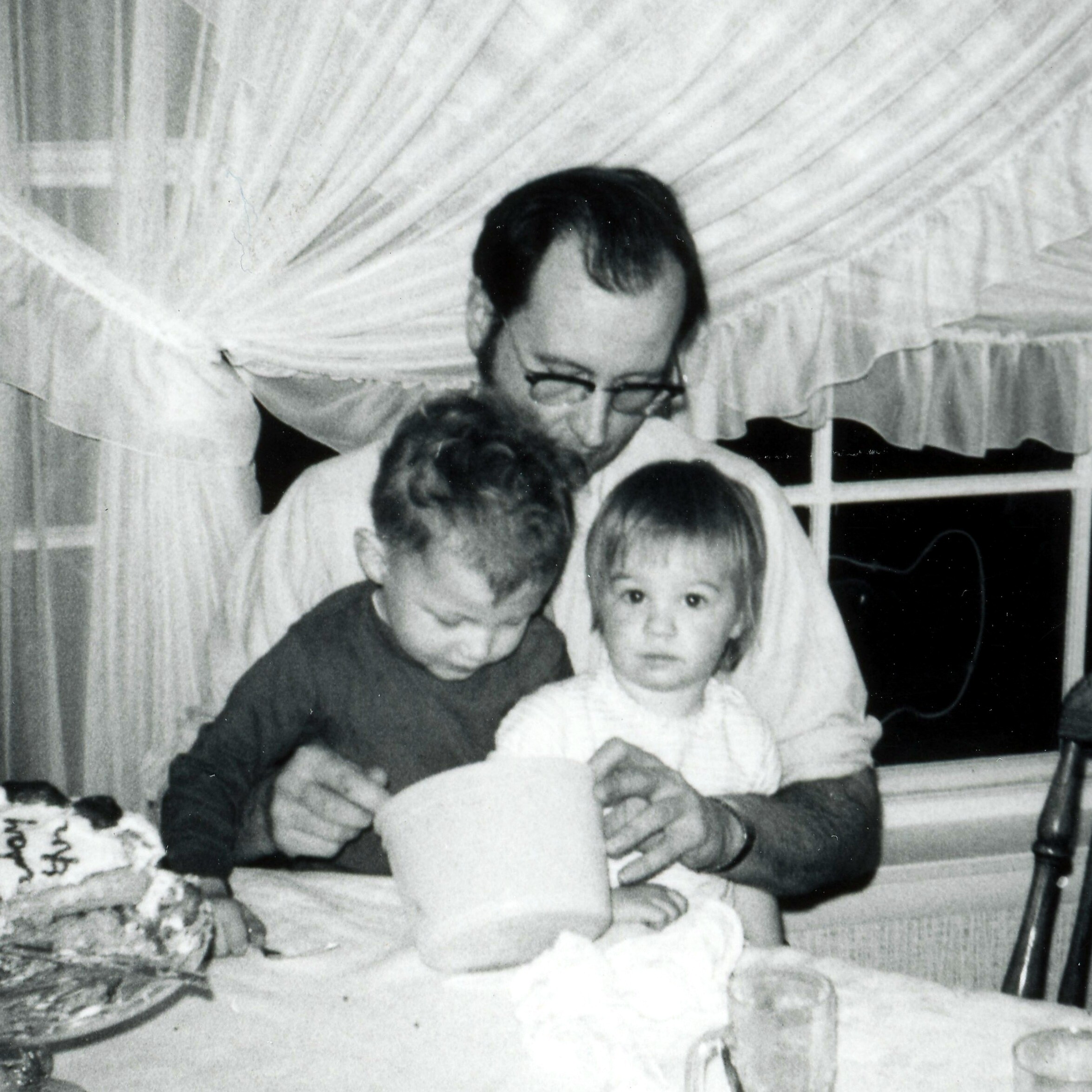 Black-and-white photo of three children gathered around a birthday cake on a table indoors.