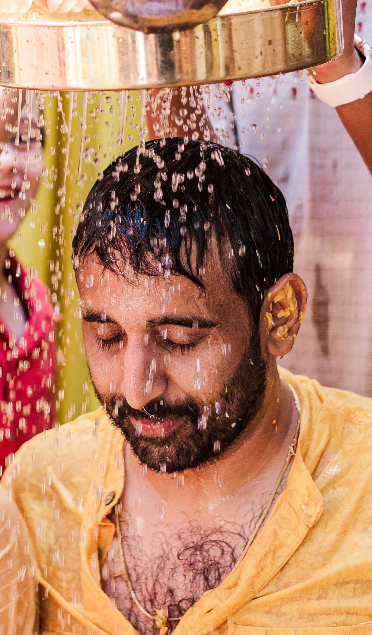 Man with eyes closed as water pours over his head from a large metal bowl during a ritual cleansing.