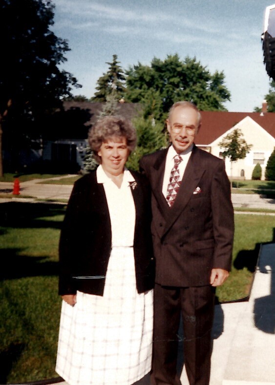 Smiling older couple standing side by side on a sunny suburban lawn; woman in a white plaid dress and black blazer, man in a dark suit and patterned tie.