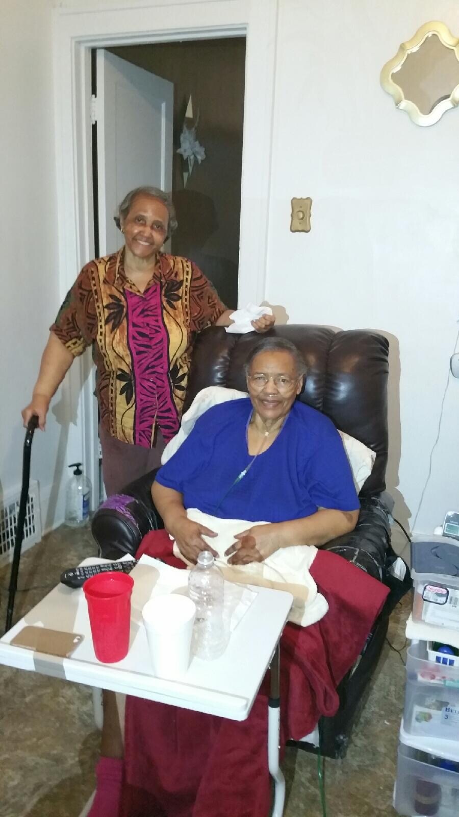 Two elderly women in a living room: one seated in a brown recliner wearing a blue top and red skirt, the other standing beside her with a tissue, wearing a colorful patterned shirt, near a small white table with cups and a water bottle in the foreground.