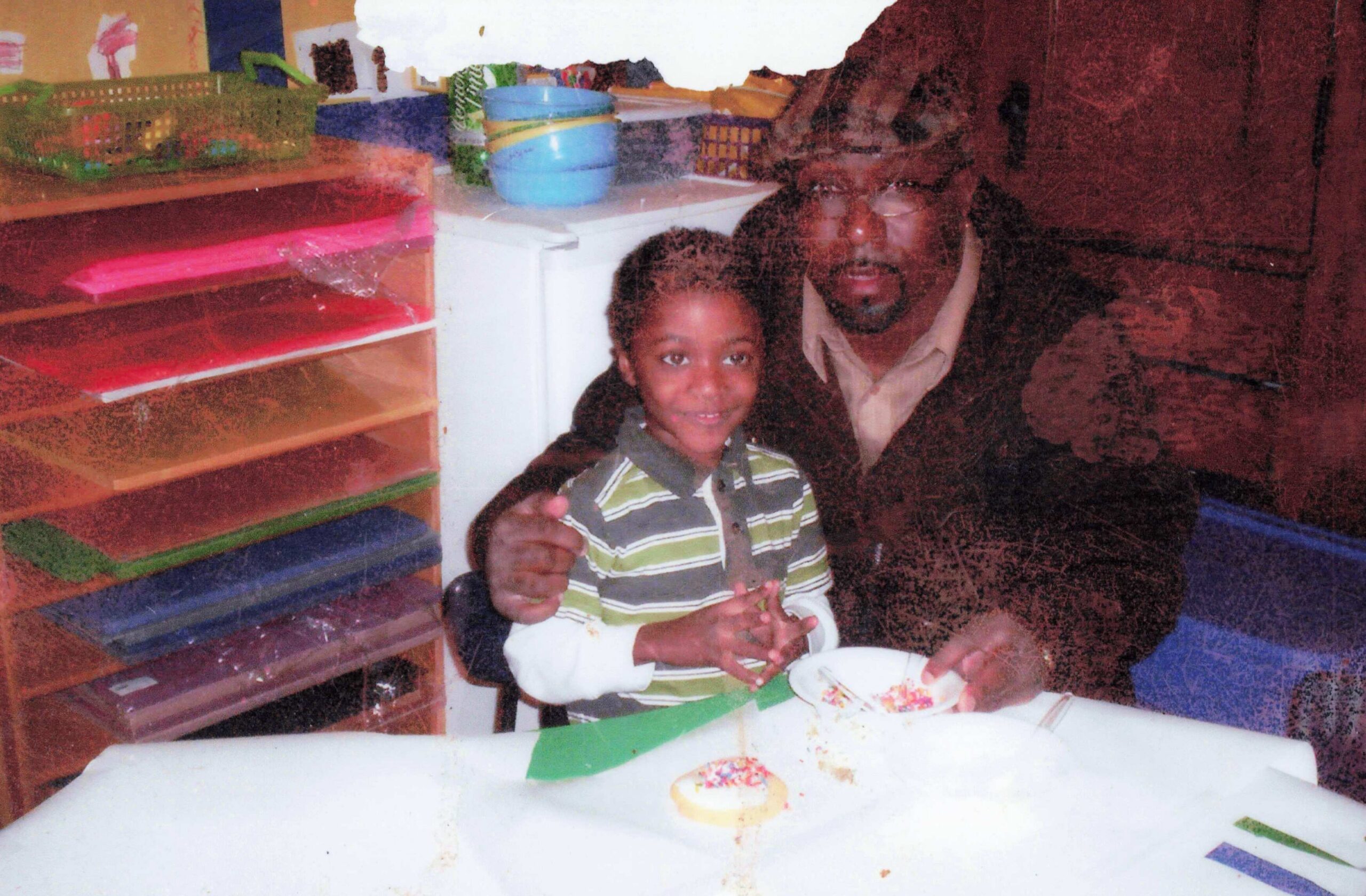 Young girl and an adult man posing together at a table with a small cake, in a colorful classroom or playroom setting.