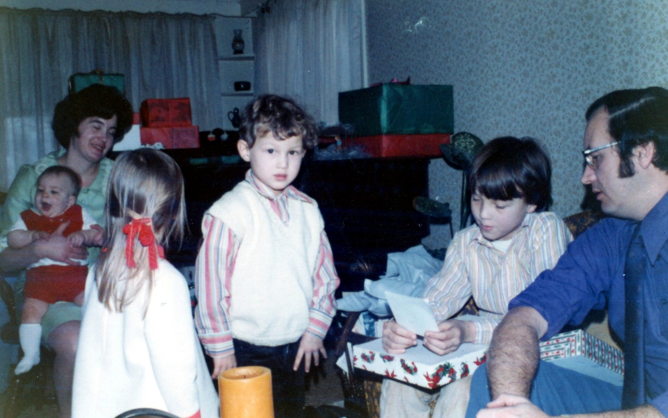 Family birthday gathering: adults and children around a table with gifts, cake, and a baby being held by an smiling woman in the background.