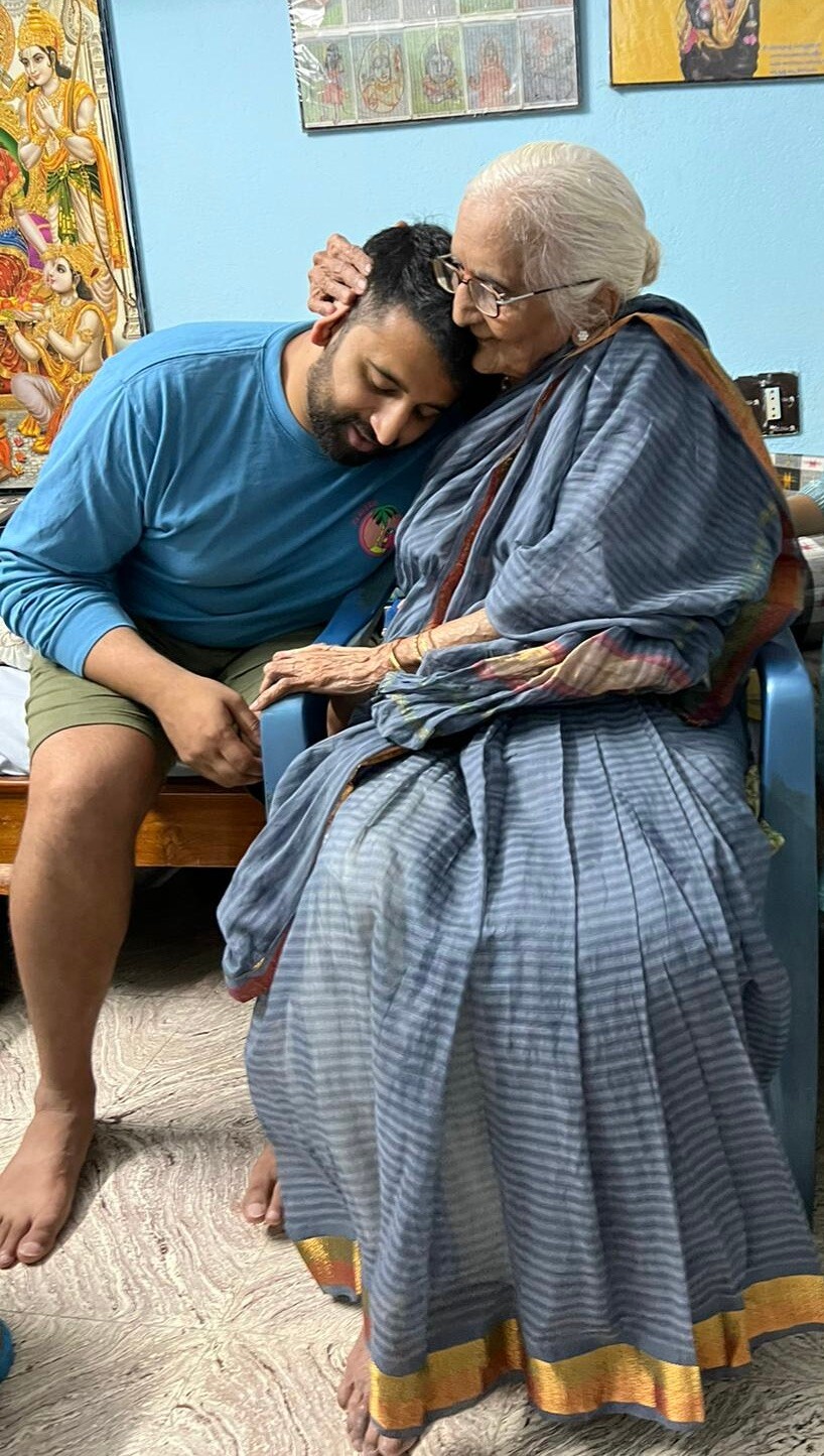 Older woman in a blue saree sits in a chair as a younger man leans his head on her shoulder, holding her hand softly.
