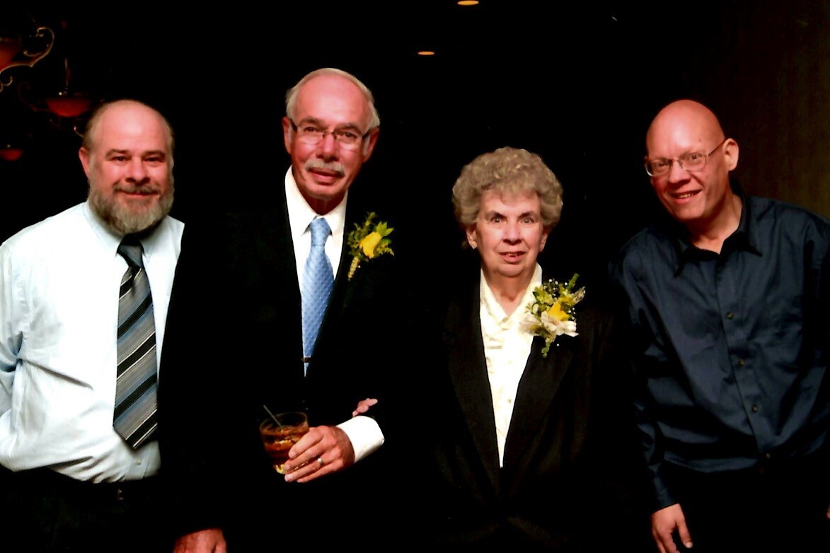 Four adults in formal attire posing at a celebration; two men wear boutonnieres and one holds a drink amid a dark background.