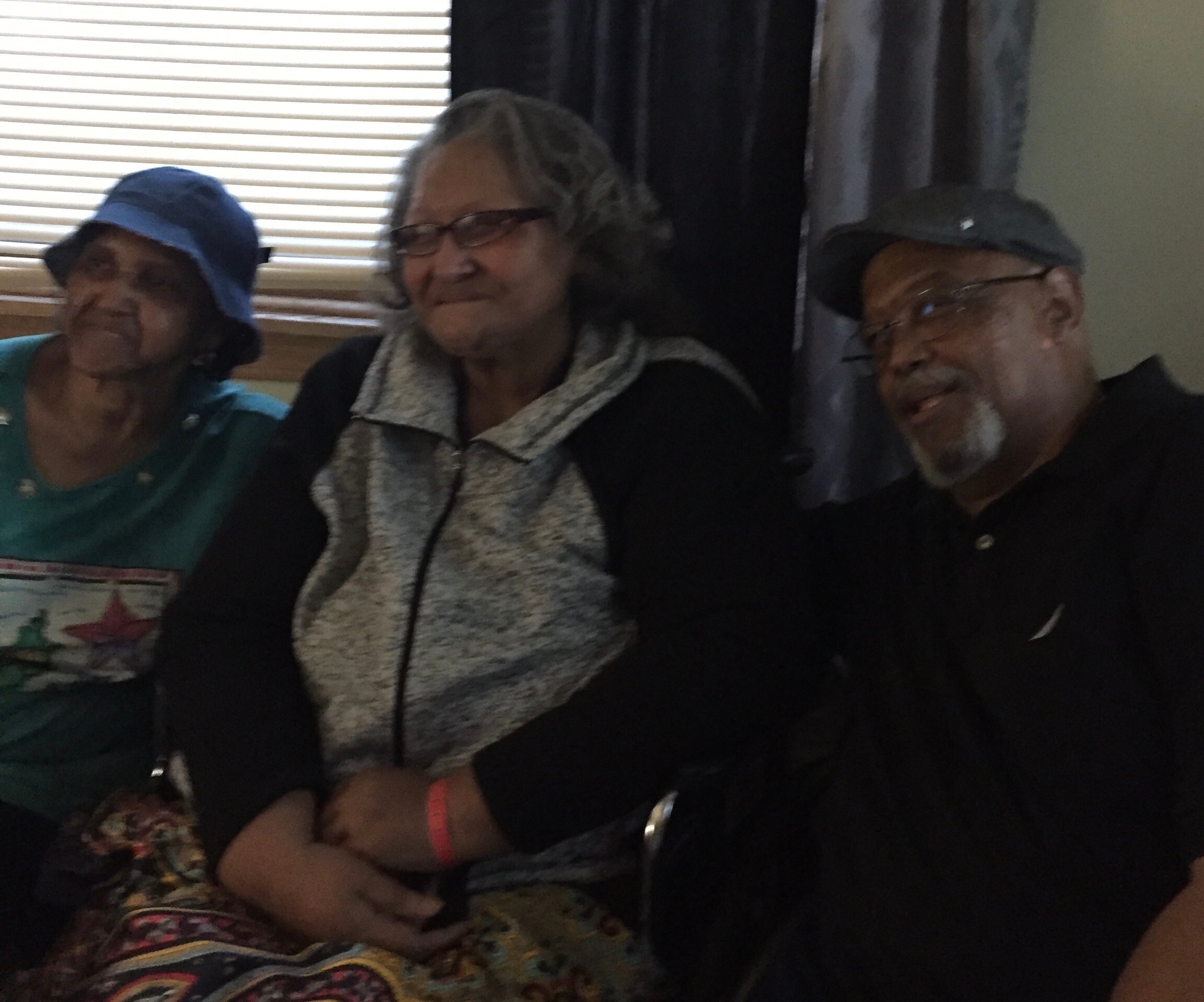 Three elderly people sit close together indoors, wearing hats and glasses, smiling softly at the camera.